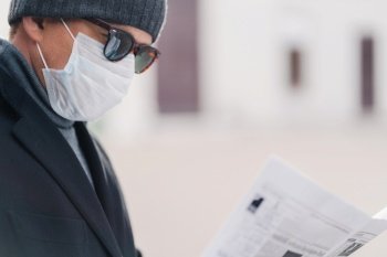 Cropped shot of male model protects against contagious disease or coronavirus, wears hygienic mask to prevent infection, respiratory illness, reads newspaper, poses outdoor in quarantined city