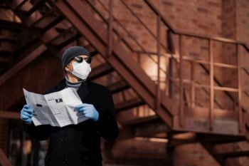 Horiontal shot of pensive man poses with newspaper, wears protective mask and rubber gloves, looks aside, thinks about self isolation, quarantine and coronavirus pandemic. People and covid 19