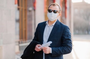 Virus, pandemic, work concept. Male manager wears formal suit and sunglasses, protective medical mask to prevent coronavirus, holds papers in hands, poses outdoor near office, waits for colleague