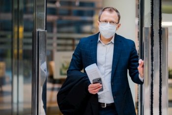 Covid-19, pandemic coronavirus concept. Office worker leaves work, wears medical face mask for infectious disease spread, dressed formally, holds newspaper and smartphone. Health and safety concept