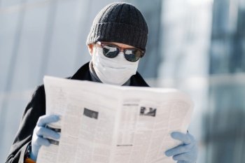 Photo of European man wears protective medical mask and gloves, walks through city outdoor, reads article about virus outbreak in different countries uses protective measures during coronavirus spread