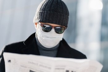 Photo of attentive man wears blak sunglasses, hat and medical mask on face during quarantine and coronavirus outbreak, reads press, finds out news from newspaper, stands against blurred background