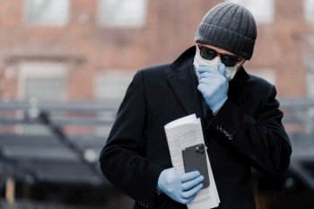 Outdoor shot of man coughs, tries not spread coronavirus infection, wears protective mask and gloves, holds newspaper and cellphone, has pneumonia, problems with breathing. Virus protection.