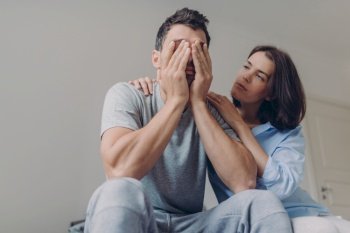 Photo of depressed male dressed casually, his beautiful woman comforts him, sit together in bedroom, have problems in family life. Affectionate wife consoles husband in difficult situation.