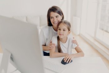 Happy affectionate mom embraces her small daughter, work together on modern computer, preapare presentation for lesson, look gladfully into monitor, spend leisure time at home, surf internet page.