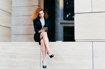 Young female model with curly hair, wearing elegant suit and high-heeled shoes, having slender legs, using modern tablet for communication. Young freelancer working outside, looking seriously