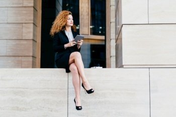 Slim businesswoman with wavy luxurious hair, having slender legs, wearing black elegant suit and shoes, holding tablet computer, looking aside while resting for minute after hard work in office