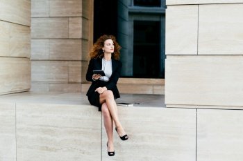 Female entrepreneur with slender legs, wearing black formal costume, sitting crossed legs, holding modern digital tablet computer, looking with pensive expression, planning her future partnership