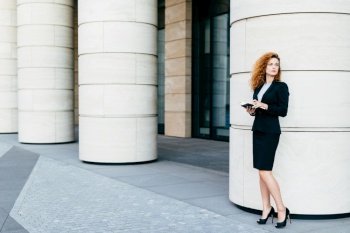 Beautiful woman with curly hair, slender legs, wearing black costume and high-heeled shoes, holding notebook in hands, arranging meeting with business partner standing near big office builduing