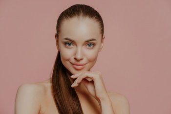 Skin care and anti aging procedures. Confident brunette young woman keeps hand under chin, has combed hair, clean fresh skin, enjoys beauty treatments, poses with naked body over pink background