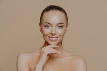 Cheerful young woman with clean glowing skin, keeps hand under chin, smiles gently, has well groomed body, combed hair, poses against beige background. Pure skin, beauty and wellbeing concept