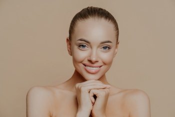 Close up shot of adult woman with fresh daily makeup, smiles toothily and keeps hands under chin, stands half naked indoor, feels refreshed after cosmetic procedures. Beauty and face care concept
