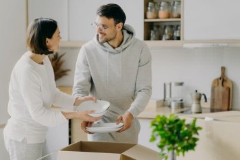 Delighted housewife and her husband unpack cardboard boxes with new tableware, carry plates, move into new apartment, look gladfully at each other, pose agaist kitchen interior. Relocation concept