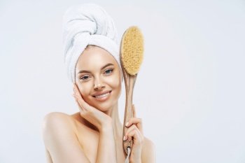 Hygiene, beauty, pampering concept. Tender smiling European woman has healthy skin, holds wooden soft massage brush for body, enjoys softness of skin after taking shower, wears wrapped towel.