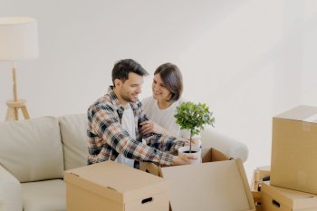 Happy couple in love settle in new home, unpack boxes with household objects, man holds potted plant, woman embraces and expresses love to husband, start new family life in modern apartment.