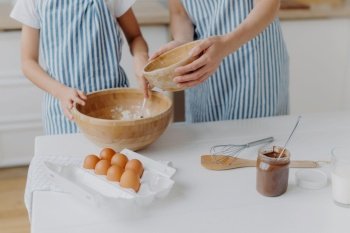 Cropped image of mothers and daughters hands mixing ingredients to prepare dough and bake tasty pastry, stand near kitchen table with eggs, melted chocolate in glass, wear striped blue aprons