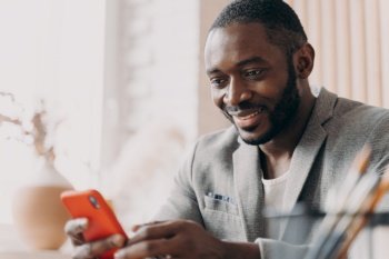 Handsome young african american businessman using mobile, chatting online while sitting at office desk, typing message and looking at phone screen with positive smile, distracted from work. Smiling african american businessman using mobile, chatting online while sitting at office desk