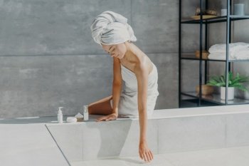 Spa, wellness and skin care concept. Serious woman wrapped in bath towel, uses cosmetic products for caring about body, going to take bath, poses on bathtub. Relaxed female at home. Spa girl