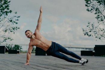 Healthy man stands in abs side plank and raises arm, does exercises outdoor, has muscular body and pleased expression. Handsome athlete trains in open air. Male couch balances on hand. Sport concept