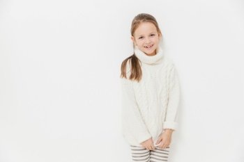 Portrait of glad satisfied girl wears warm white knitted sweater, has joyful expression, stands against studio background with blank copy space. Adorable female child enjoys leisure time. Monochrome
