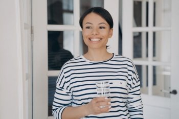 Smiling female holds glass with eco mineral clean water, standing at home. Happy woman enjoying fresh pure potable aqua. Healthy lifestyle habit, wellness concept. Water filter advertisement.. Happy female holding glass of clean water. Healthy lifestyle, wellness. Water filter advertising