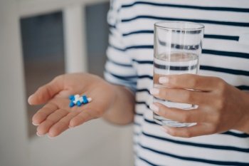 Hand of european woman is holding white and blue capsules and glass of water. Lady is going to take drugs. Therapy with medicine. Supplements and vitamins, pharmacology, influenza curation.. Hand of woman is holding white and blue capsules and glass of water. Lady is going to take drugs.