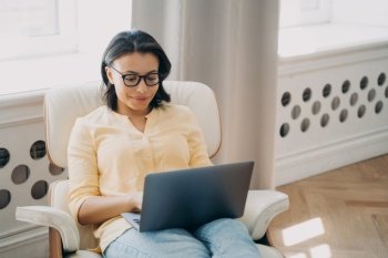 Young happy caucasian woman is studying online. Lady is working on laptop sitting in an arm chair. Lectures or webinars, internet conference. Remote self education online on quarantine, autodidact.. Young happy caucasian woman studying online on laptop. Remote self education online on quarantine.