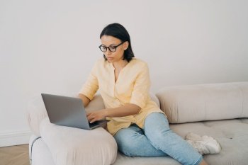 Concentrated woman is student, working laptop at home on couch. Young mixed race freelancer has remote work. Lady is studying online on quarantine. Business woman or freelancer is doing job from home.. Concentrated woman is student, business lady or freelancer, working online on quarantine.