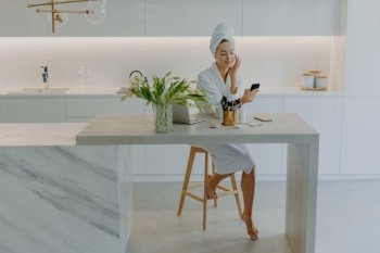 Beautiful young female model poses in modern apartment wears bathrobe and towel on head applies facial cream undergoes beauty procedures uses cosmetic tools for skin care. Pampering wellness