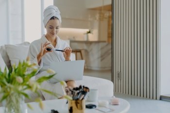 Lovely young Caucasian woman wears bathrobe and towel on head holds cosmetic brush in front of laptop computer gives tutorail lesson how to do makeup sits at comfortable sofa in modern apartment