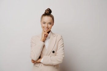 Professional attractive business lady in suit looking at camera calmly confidently, thinking about company and next move in carrier while standing a in front of grey background, copy space for text. Professional attractive business lady keeps hand under chin