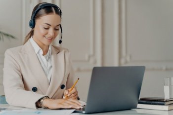 Portrait of friendly female CEO wearing headset with microphone taking part in web conference on laptop computer, positive young businesswoman in formal suit writing notes while sitting at workplace. Positive young businesswoman in headset taking part in web conference