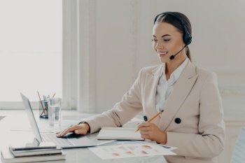 Side view of young smiling business woman wearing taking part in web conference, looking at laptop screen, call center operator consulting customer while sitting behind work desk in modern office. Call center operator wearing headset talking with client