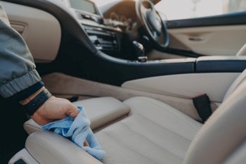 Cropped shot of man cleaning car interior, wiping fabric passenger seat with microfiber cloth, removing dirt and dust in modern vehicle salon with rag. Car wash concept. Man cleaning car interior with microfiber cloth at carwash service