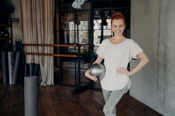 Young smiling pilates instructor with ginger hair standing in fitness studio or gym with her hand on waist, holding small silver fitball and looking into camera positively, dressed in active wear. Smiling fit female posing with pilates ball in fitness studio
