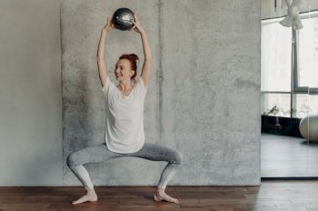 Full leghth shot of slim happy ginger female performing barre exercises with small fitball lifted up over head by two hands and sitting in deep plie in fitness studio, dressed in activewear. Slim pretty ginger woman enjoying barre exercises with small fitball