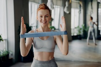 Portrait of young sporty happy redhead woman wearing white top and gray leggings performs exercises for muscles of hands during workout with resistance band. Fitness instructor during sports class. Portrait of young sporty redhead woman in sportswear with resistance rubber band