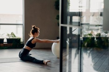 View through open glass door of flexible redhead slender female in active wear sitting in twine on mat with hands laying on big fitball while performing stretching exercises in gym or fitness studio. Young redhead slender woman sitting in twine with fitball, doing stretching exercises