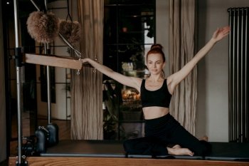 Slim young female pilates instructor sitting on trapeze with arms outstretched to both sides in relaxed position doing stretching exercises in gym on cadillac reformer. Sport and fitness concept. Flexible woman sitting on trapeze with arms outstreched to both sides