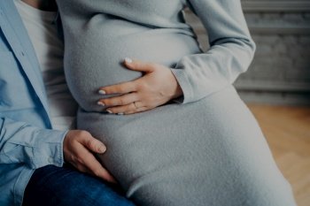 Cropped shot of unrecognizable parents await for baby. Pregnant woman with big belly poses on husbands hands feels child movement in abdomen. Parenthood concept
