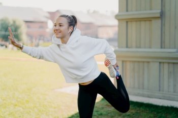 Outdoor shot of pretty young Eruopean woman in sportswear does warm up exercises outdoors stretches legs before run looks into distance with happy expression listens music in wireless earphones
