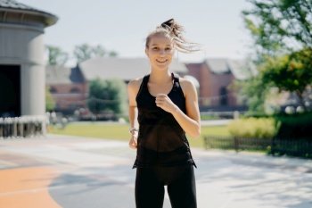 Happy energetic sportswoman runs on stadium has pony tail floating on wind dressed in activewear being active runner has positive smile enjoys regular morning trainings motivates you for sport