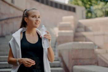 Athlete woman with pony tail feels thirsty after cardio training drinks water from bottle holds refreshig drink stays hydrated poses outdoors dressed in active wear concentrated into distance
