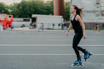 Energetic slim young smiling woman rollerblades on city road enjoys spending free time actively moves quickly has dark hair floating on wind leads healthy lifestyle. Active weekends. Outdoor photo