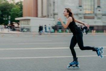 Outdoor photo of active dark haired woman in good physical shape enjoys extreme sport rides on rollers along road at busy street relaxes during weekend in open air. Healthy lifestyle concept
