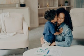 Cute mixed race boy little kid gently hugs his happy mother sitting on floor by table with colored pencils on it, surrounded by light colored furniture in living room. Leisure time with family concept. Cute little kid with curly hair gently hugging his smiling mother sitting on floor in living room