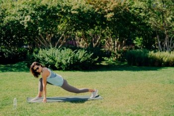 Outdoor sport concept. Healthy fit energetic young woman does fitness exercises on fitness mat dressed in active wear drinks fresh water wears sunglasses poses on green grass. Morning workout.