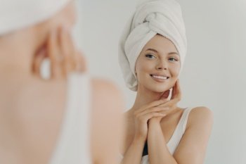 Beautiful young woman after shower looks in mirror gently touching soft healthy glowing face skin, happy millennial female applying beauty products during daily facial procedures, skincare concept. Beautiful young woman after shower looking in mirror in bathroom