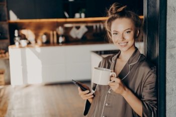 Young positive business woman wears silk brown pajama smiling while holding smartphone in hand and drinking coffee after waking up in morning at home before going to work, standing in bedroom. Portrait of young positive woman holding smartphone in hand and drinking coffee while standing in bedroom