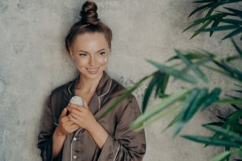 Image of caucasian cheerful woman in silk brown pajama smiling while applying face cream isolated over concrete wall background. Daily routine procedure for rejuvenation and care of skin. Young caucasian attractive woman in silk brown pajama doing skin care procedure at home